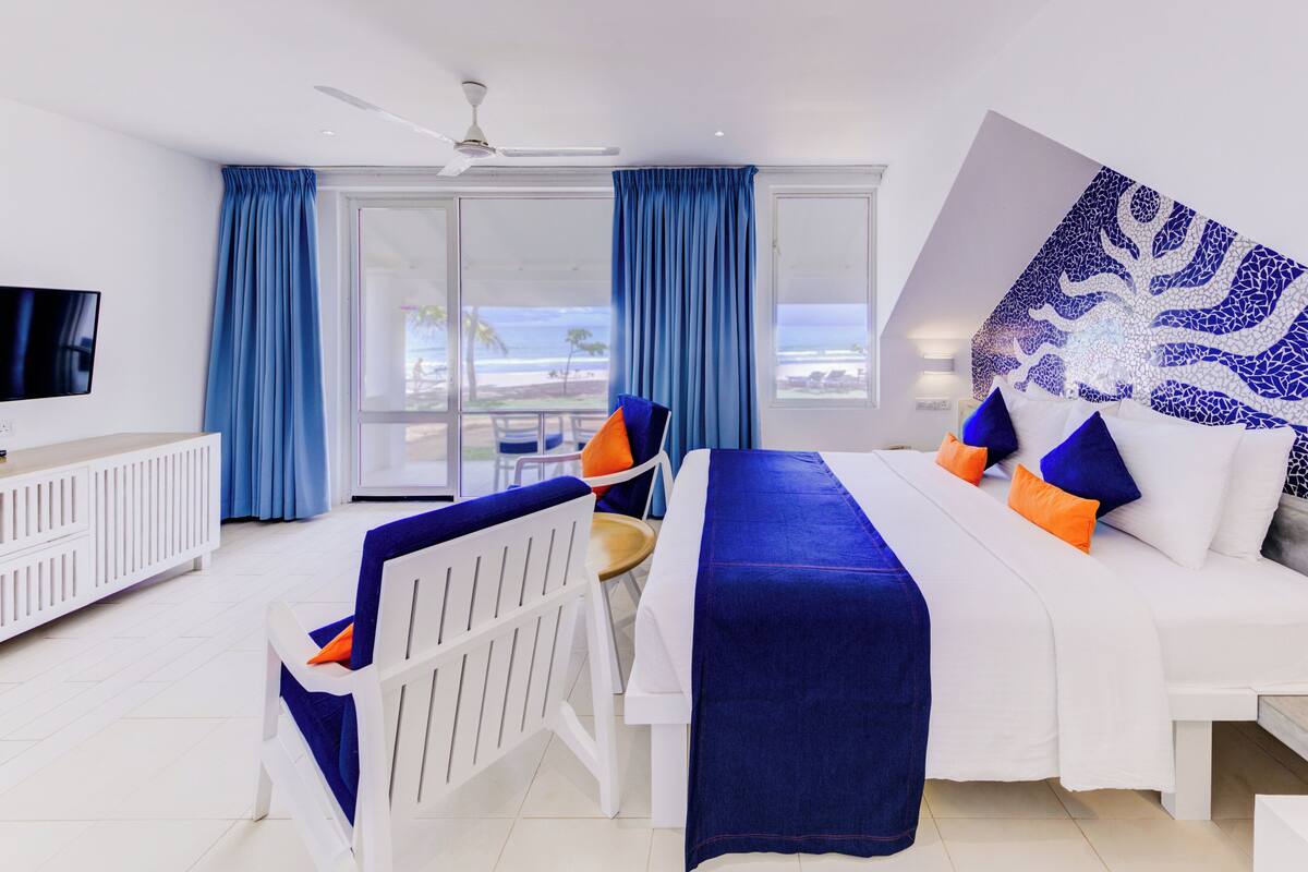 A hotel room with a bed, a TV, a desk, and a view of the beach.