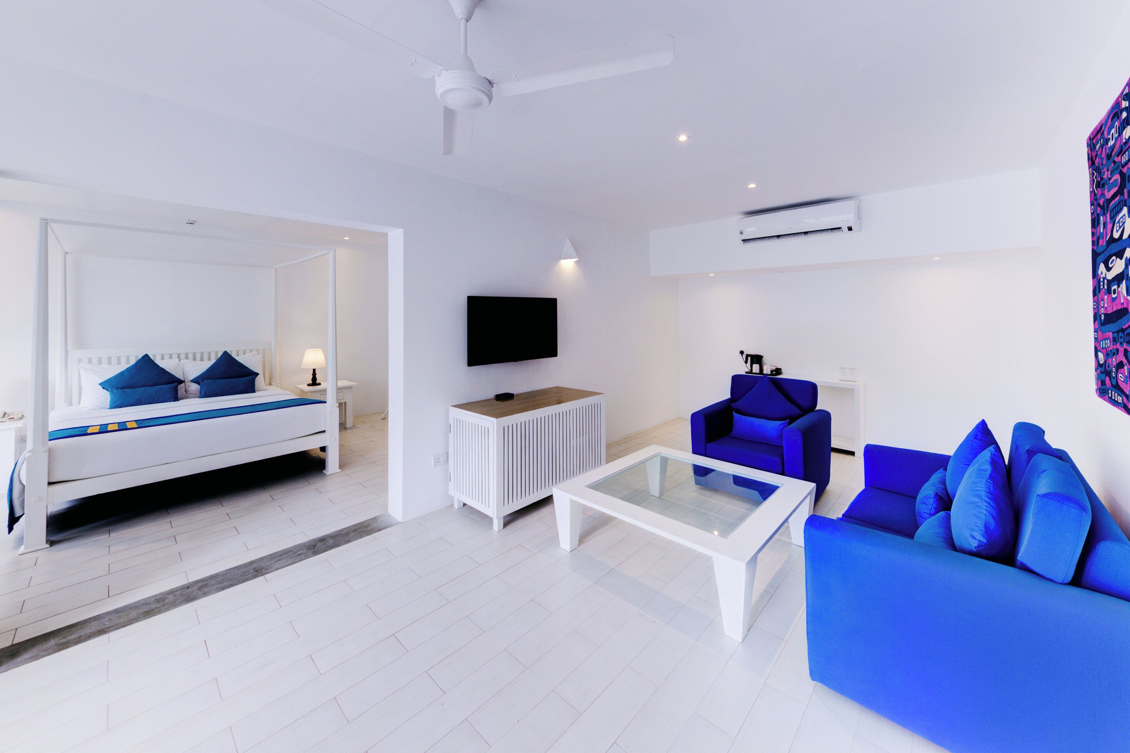 A modern hotel room with a bed, a blue sofa, a coffee table, a TV, and a ceiling fan.