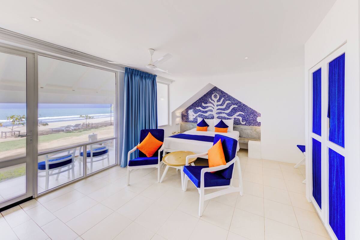 A hotel room with a balcony, a bed with blue and orange bedding, and a view of the beach.