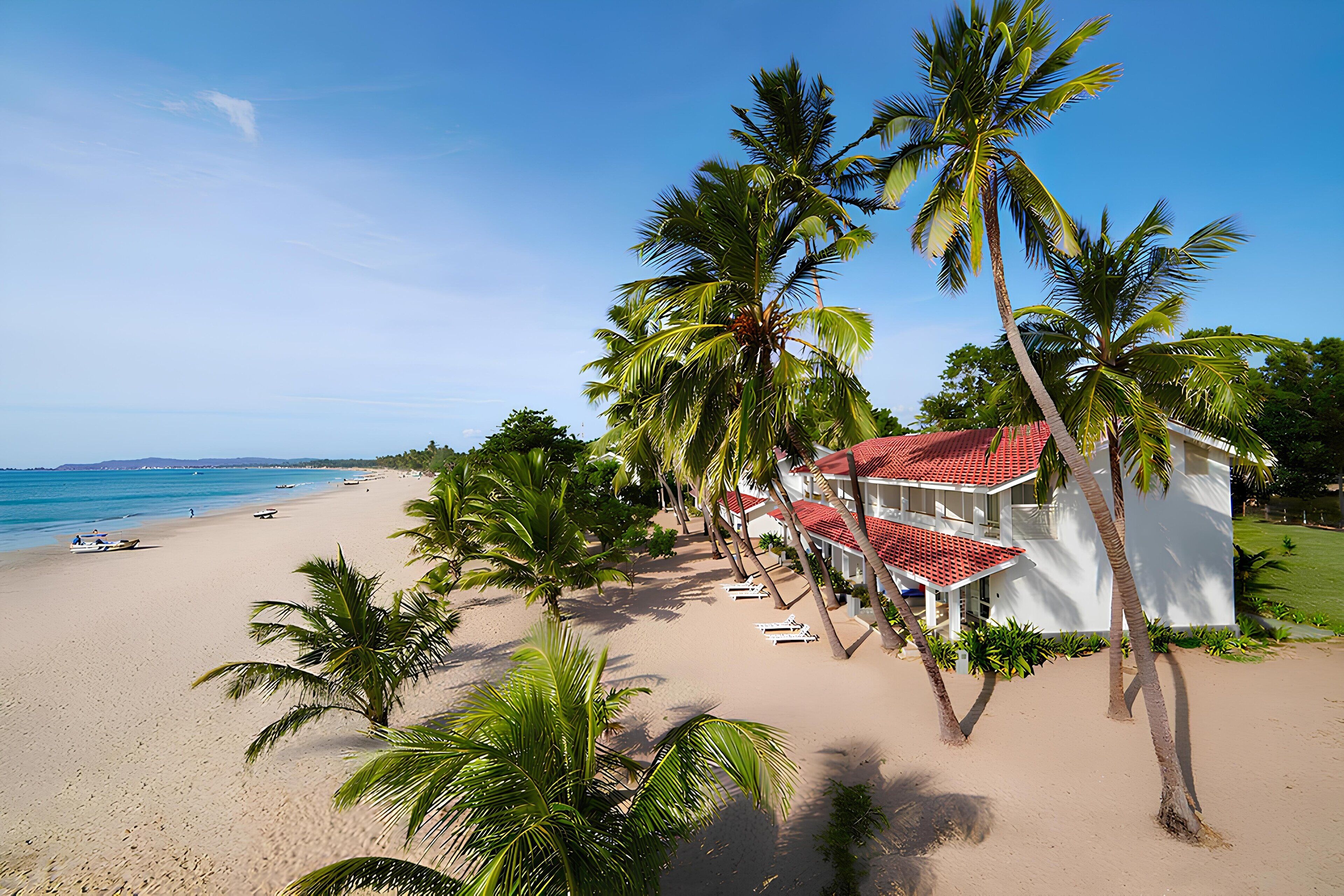 A beachfront hotel with a red roof, palm trees, and a sandy beach.
