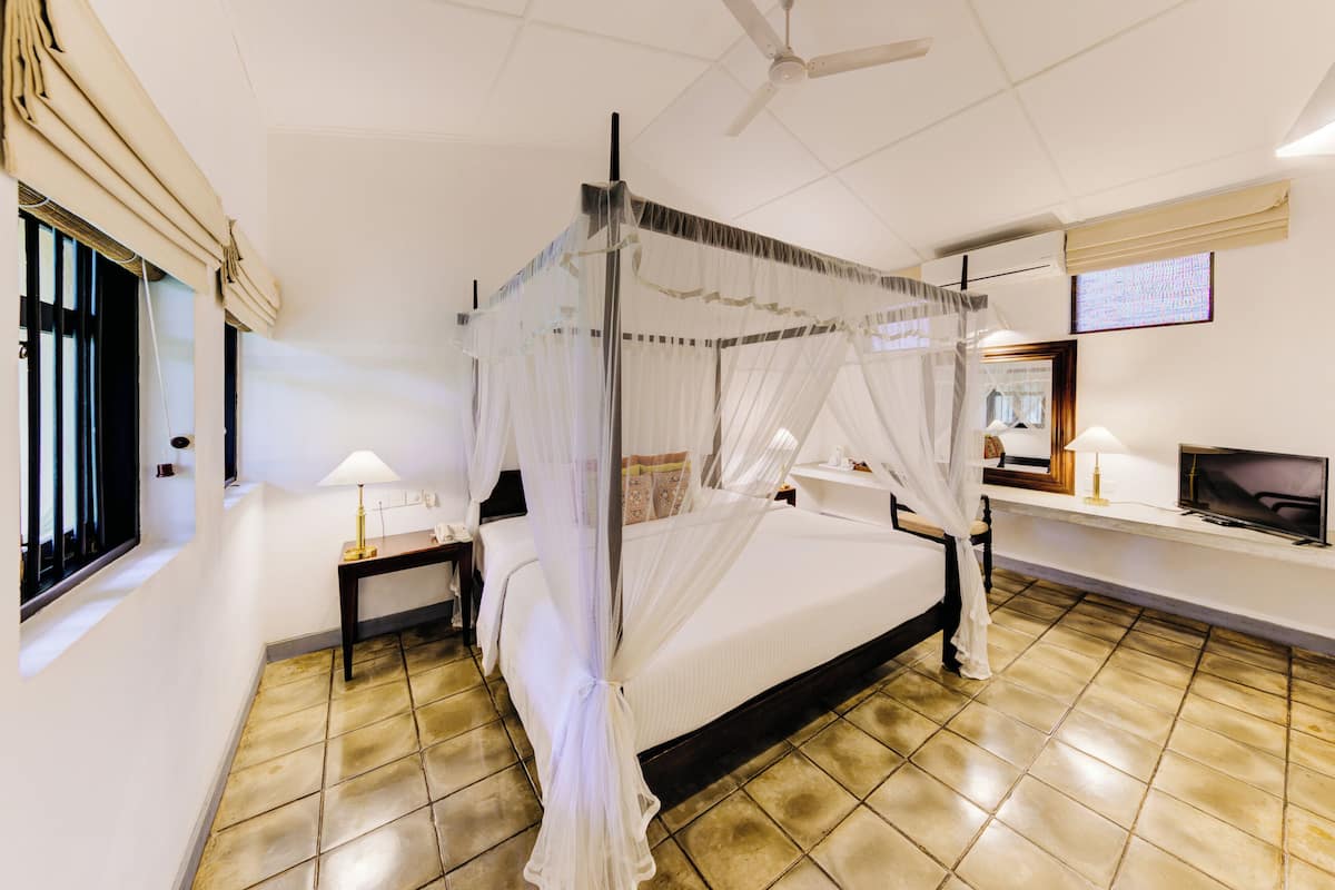 Un lit à baldaquin, une table de chevet avec une lampe, un téléviseur posé sur un meuble et une fenêtre avec des stores.