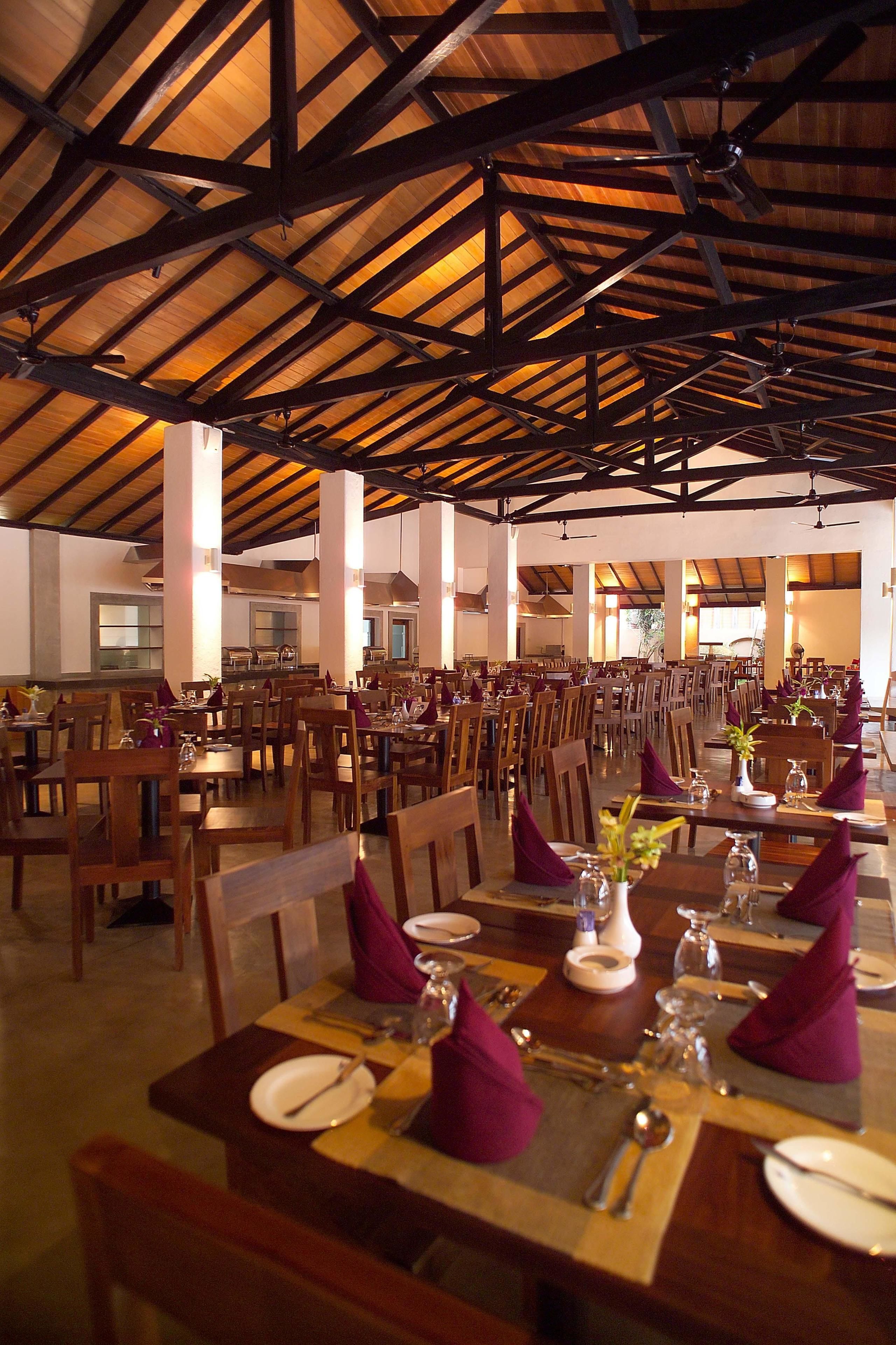 A spacious dining area with wooden tables and chairs, arranged in rows, under a high wooden ceiling with exposed beams.