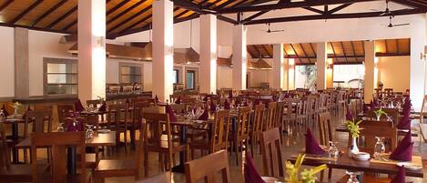 A spacious dining area with wooden tables and chairs, arranged in rows, under a high wooden ceiling with exposed beams.