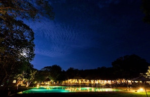 Vista nocturna de un hotel con piscina, mobiliario exterior y árboles.