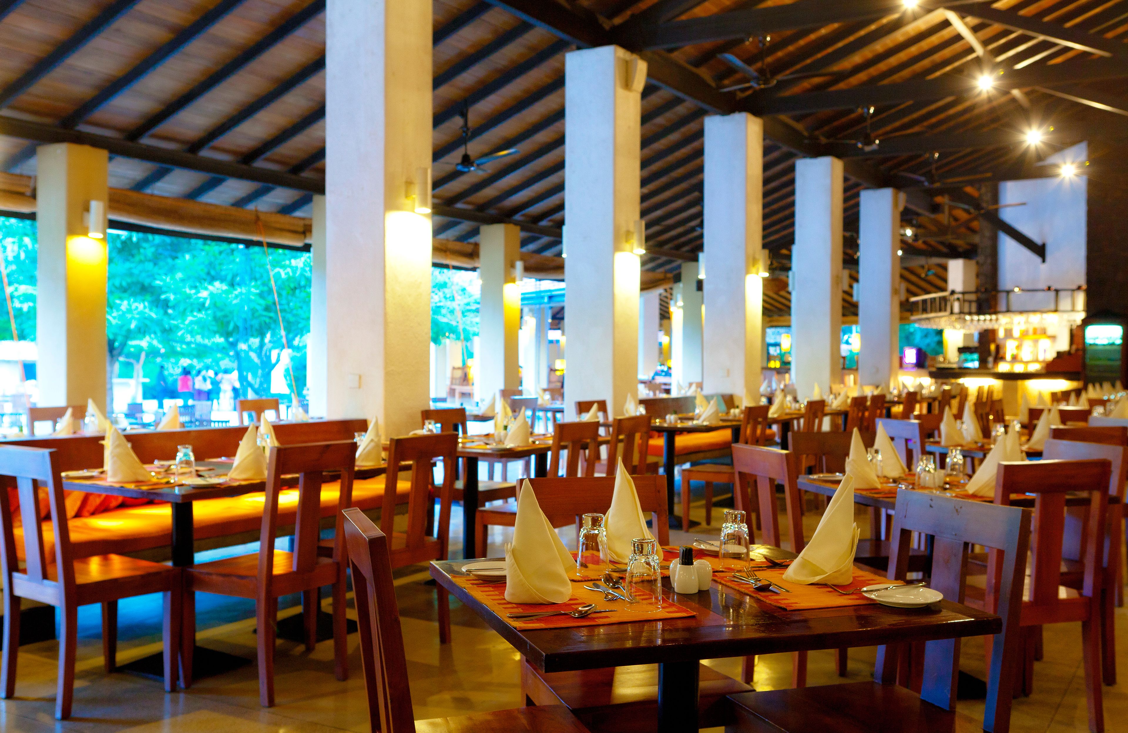 A restaurant with wooden tables and chairs, each set with a napkin, salt, pepper, and a glass.
