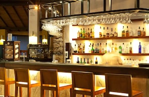 Zona de bar con sillas de madera, una estantería iluminada con varias botellas y un colgador para copas de vino.