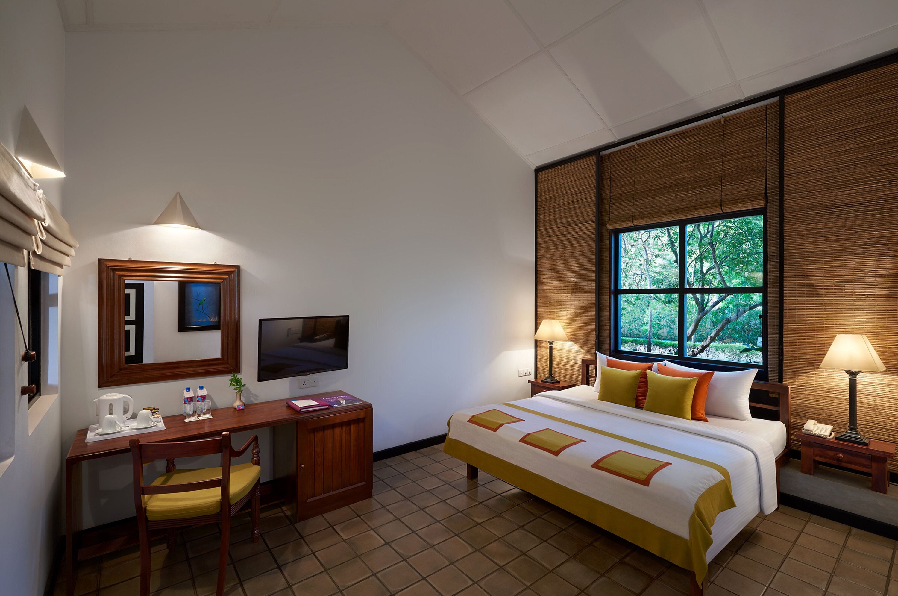 A hotel room with a bed, desk, chair, TV, and window with a view of trees.