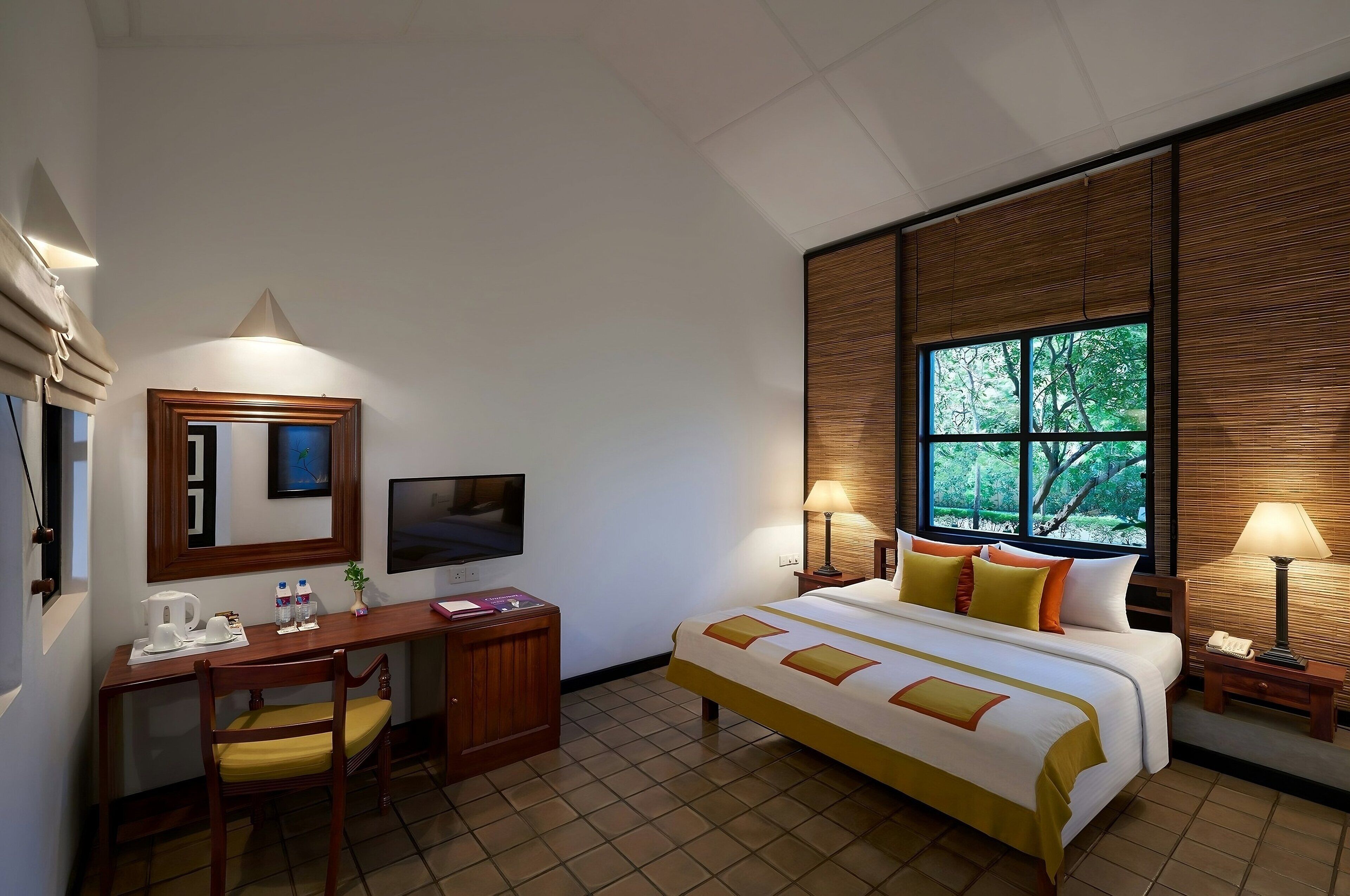 Une chambre d’hôtel équipée d’un lit, d’un bureau, d’une chaise, d’une télévision et d’une fenêtre donnant sur un paysage arboré.
