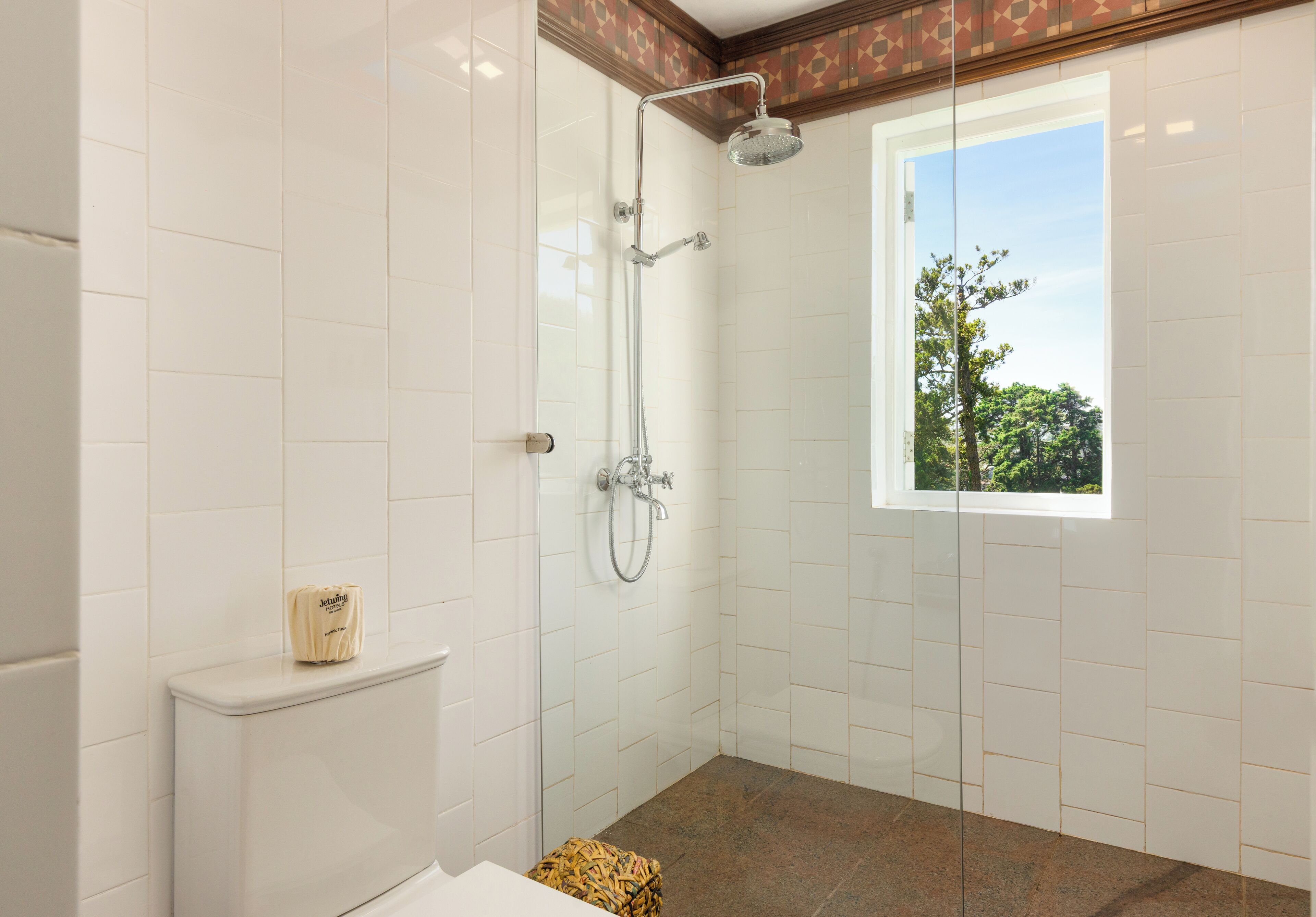 Ein modernes Badezimmer mit weißen Fliesen, einer Dusche, einer Toilette und einem Fenster mit Blick auf Bäume.