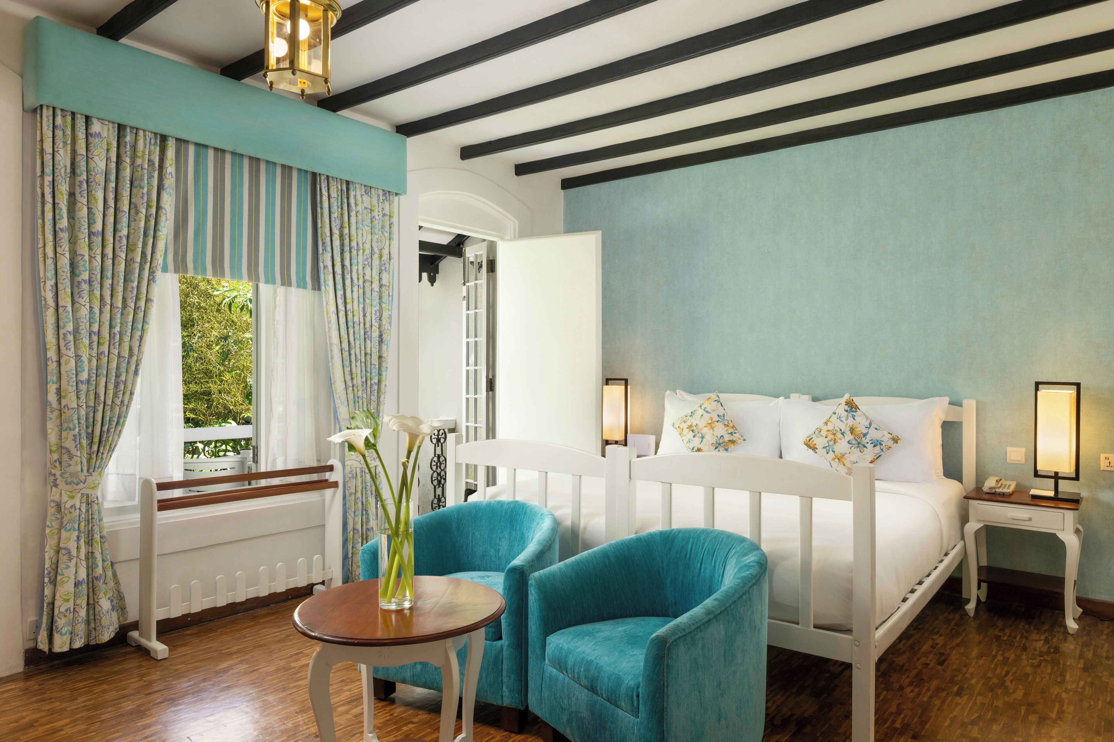A bedroom with a bed, two blue chairs, a small table, a nightstand, and a window with curtains.