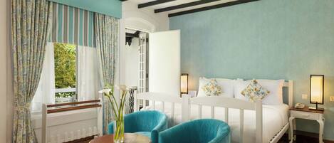 A bedroom with a bed, two blue chairs, a small table, a nightstand, and a window with curtains.