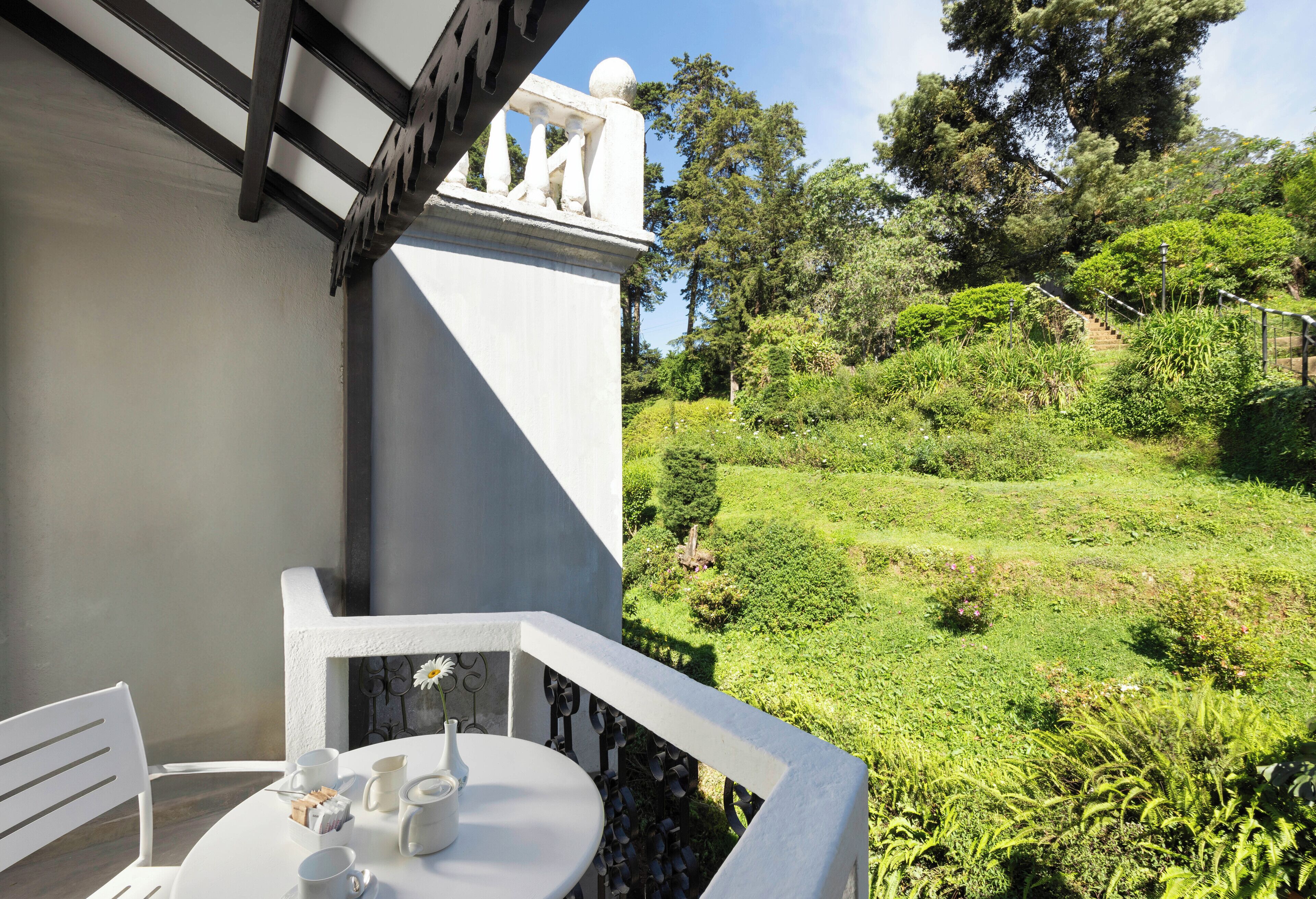 Un balcón con una mesa y sillas blancas, con vista a un jardín exuberante y una escalera que sube una colina.