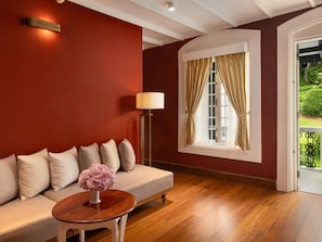 Ein Zimmer mit roter Wand, Holzboden, einer Couch mit Kissen, einem Tisch mit einer Vase, einer Stehlampe und einem Fenster mit Vorhängen.