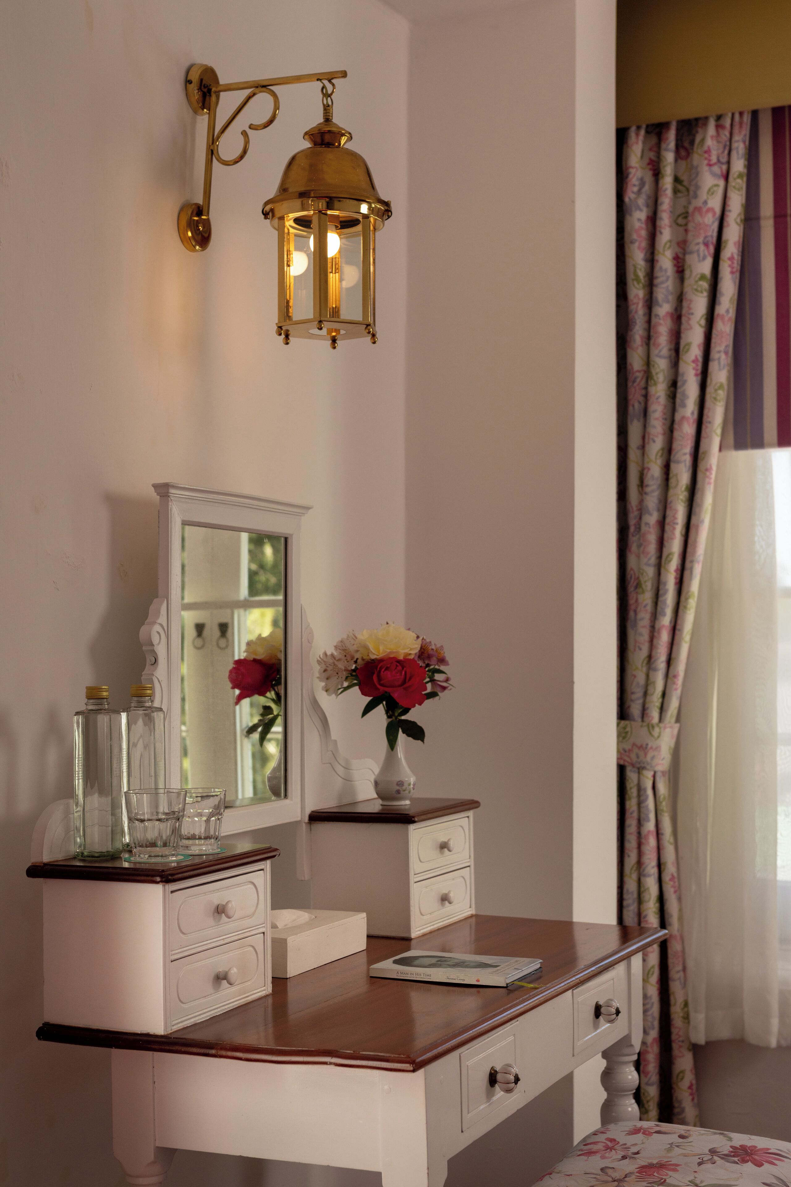 A bedroom with a wooden dresser, a mirror, a vase with flowers, and a wall-mounted lamp.