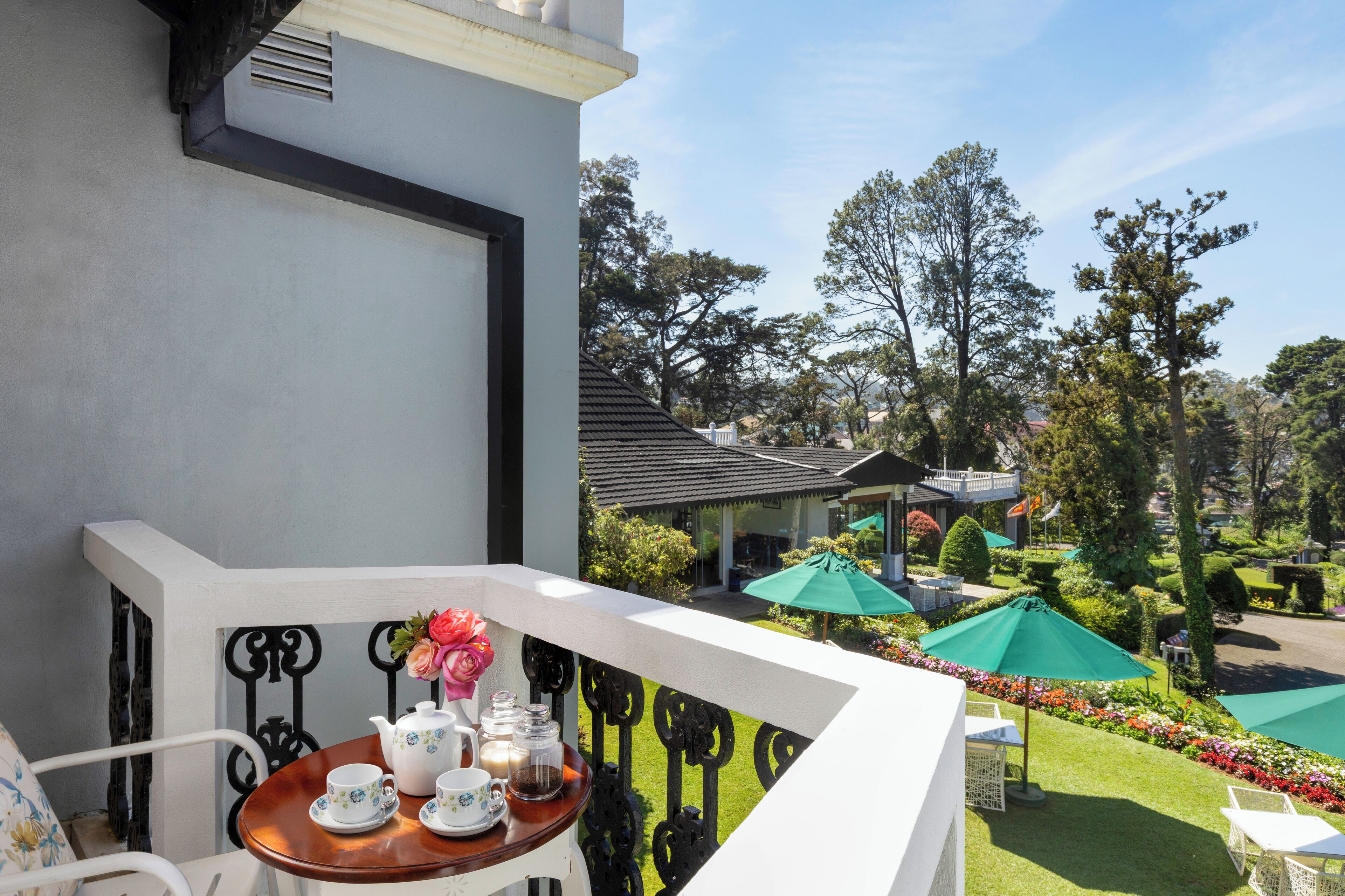 Un balcon avec une table dressée pour le thé, donnant sur un jardin agrémenté de parasols et sur un bâtiment en arrière-plan.