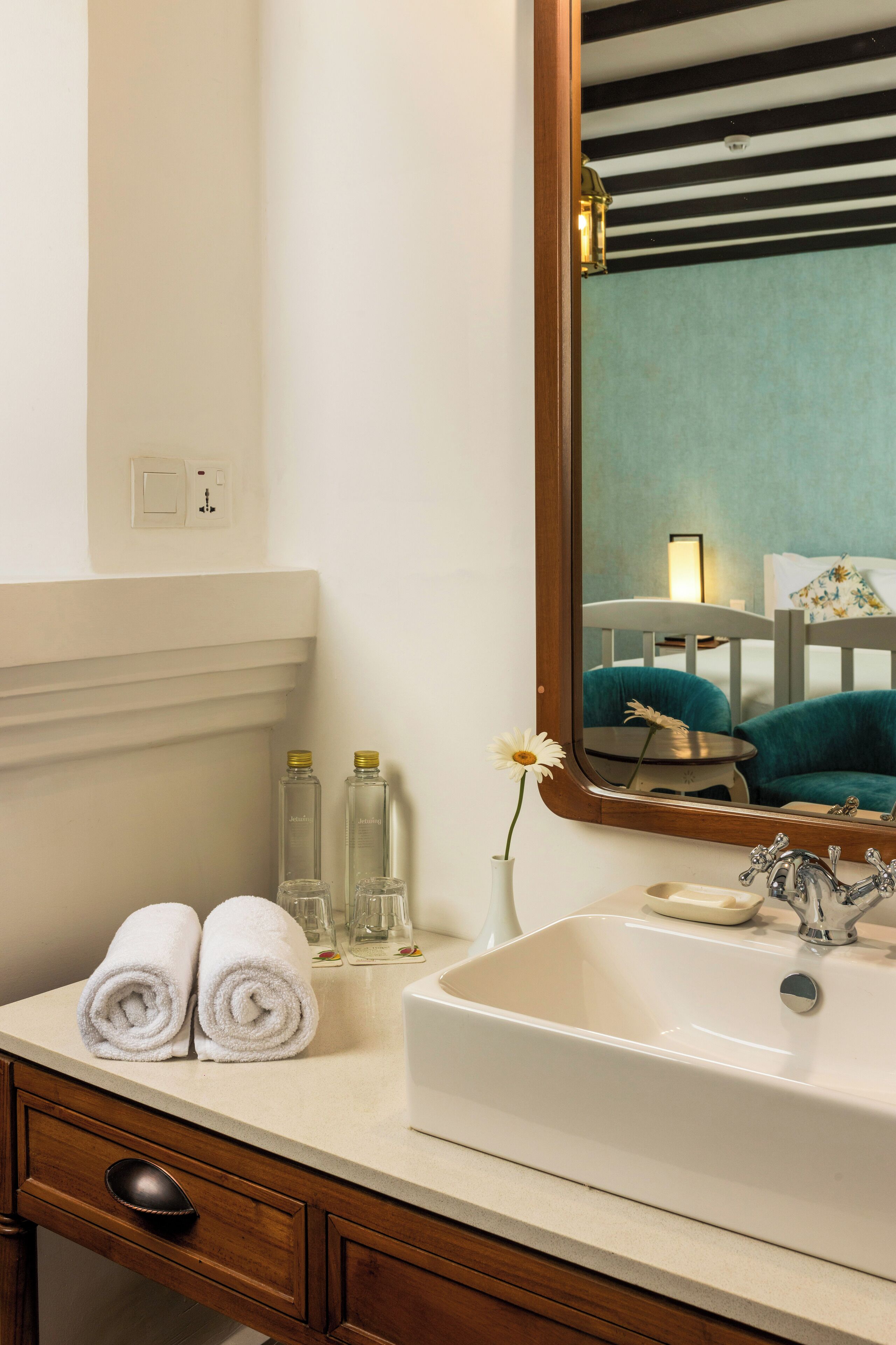Une salle de bain avec un lavabo blanc, deux serviettes roulées, un vase avec une fleur et un miroir qui reflète une chaise turquoise et une table.