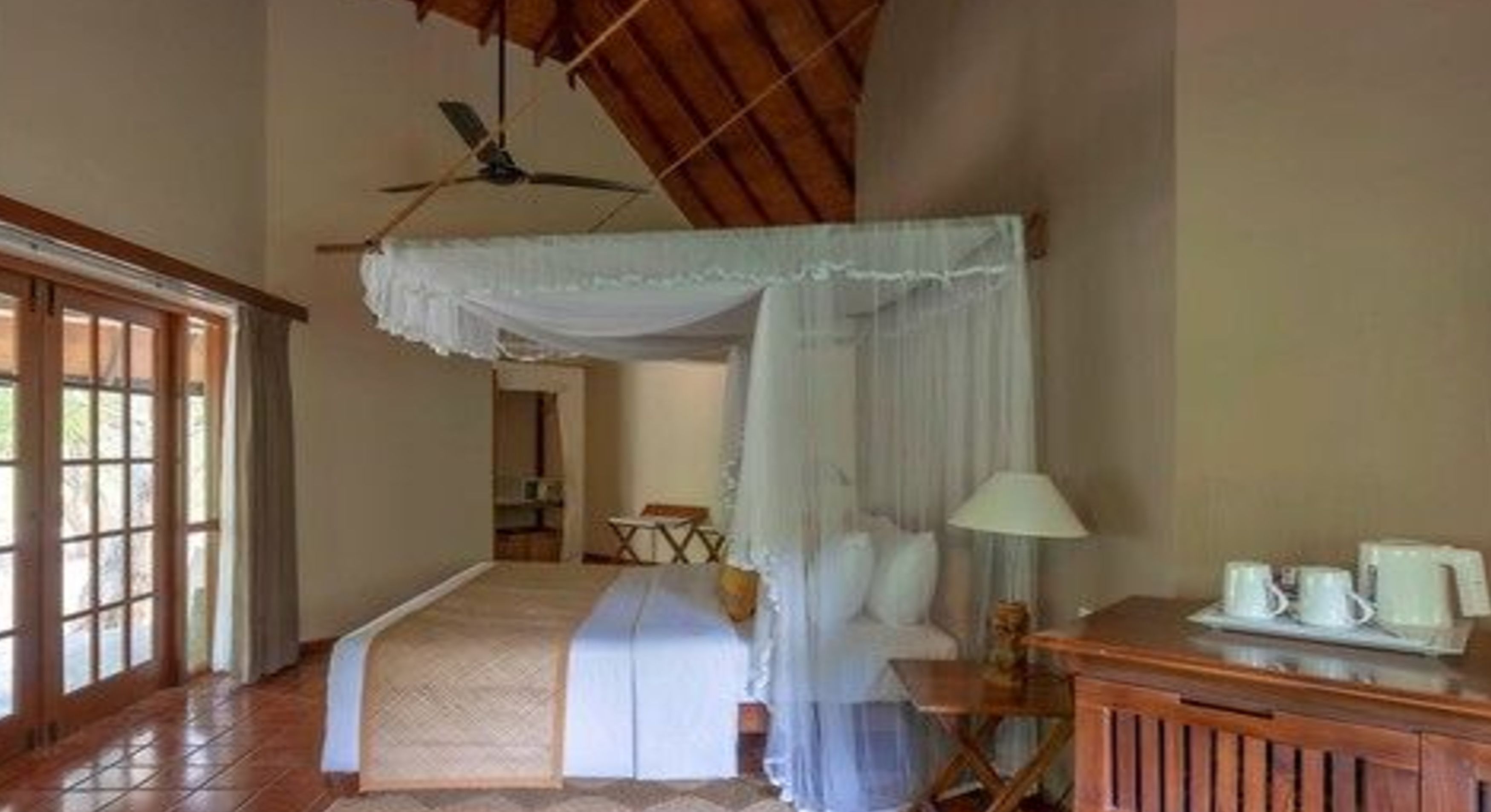 Quarto com cama sob um dossel, escrivaninha de madeira com abajur e vista para uma varanda através de uma janela.