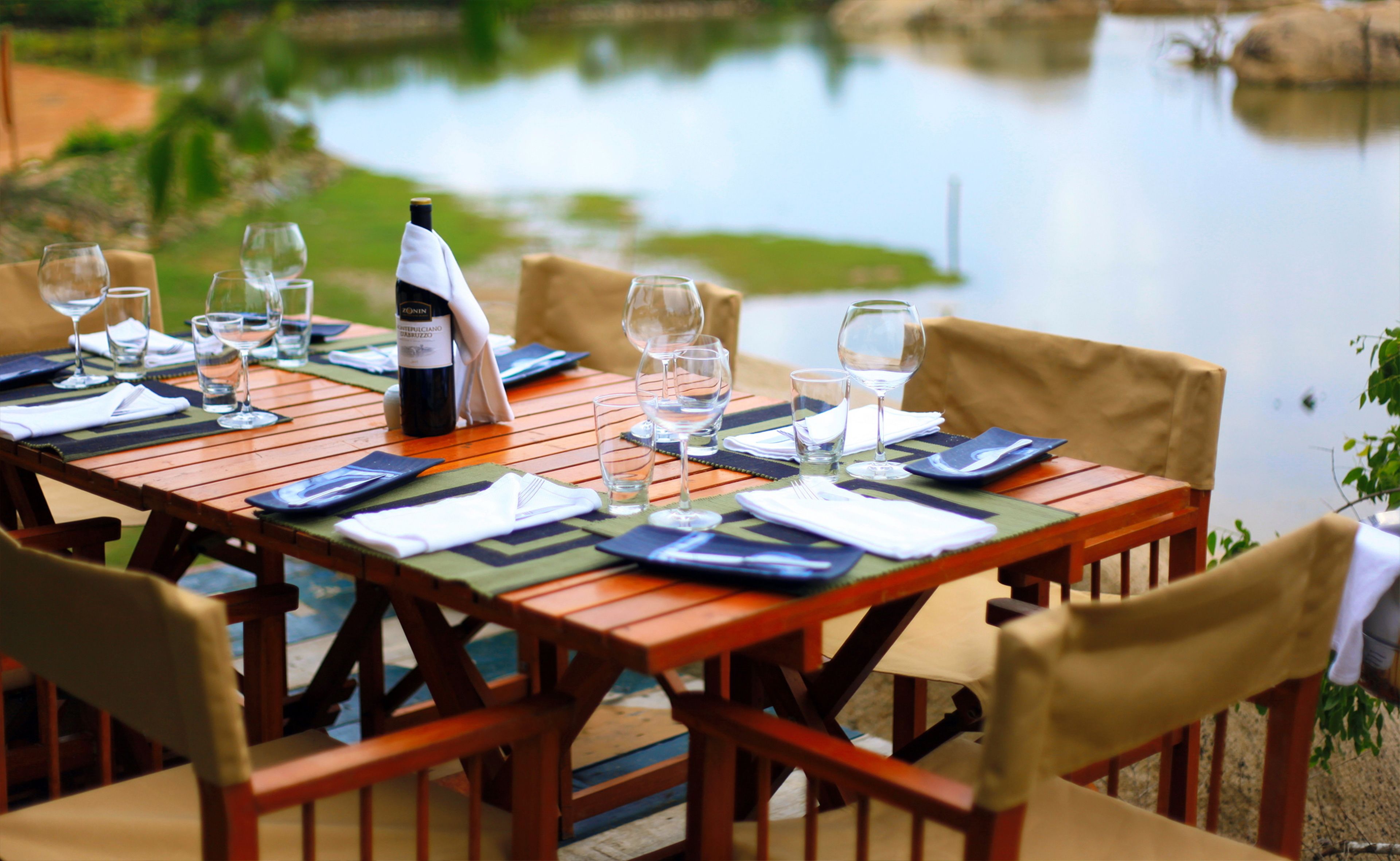 Una mesa de comedor dispuesta con copas de vino, platos y una botella de vino, con vista a un cuerpo de agua tranquilo.