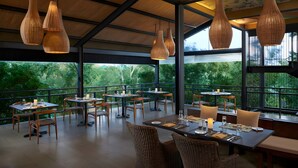 A restaurant with tables set for dining, hanging wicker pendant lights, and large windows offering a view of greenery. - Cinnamon Wild Yala (Kirinda)