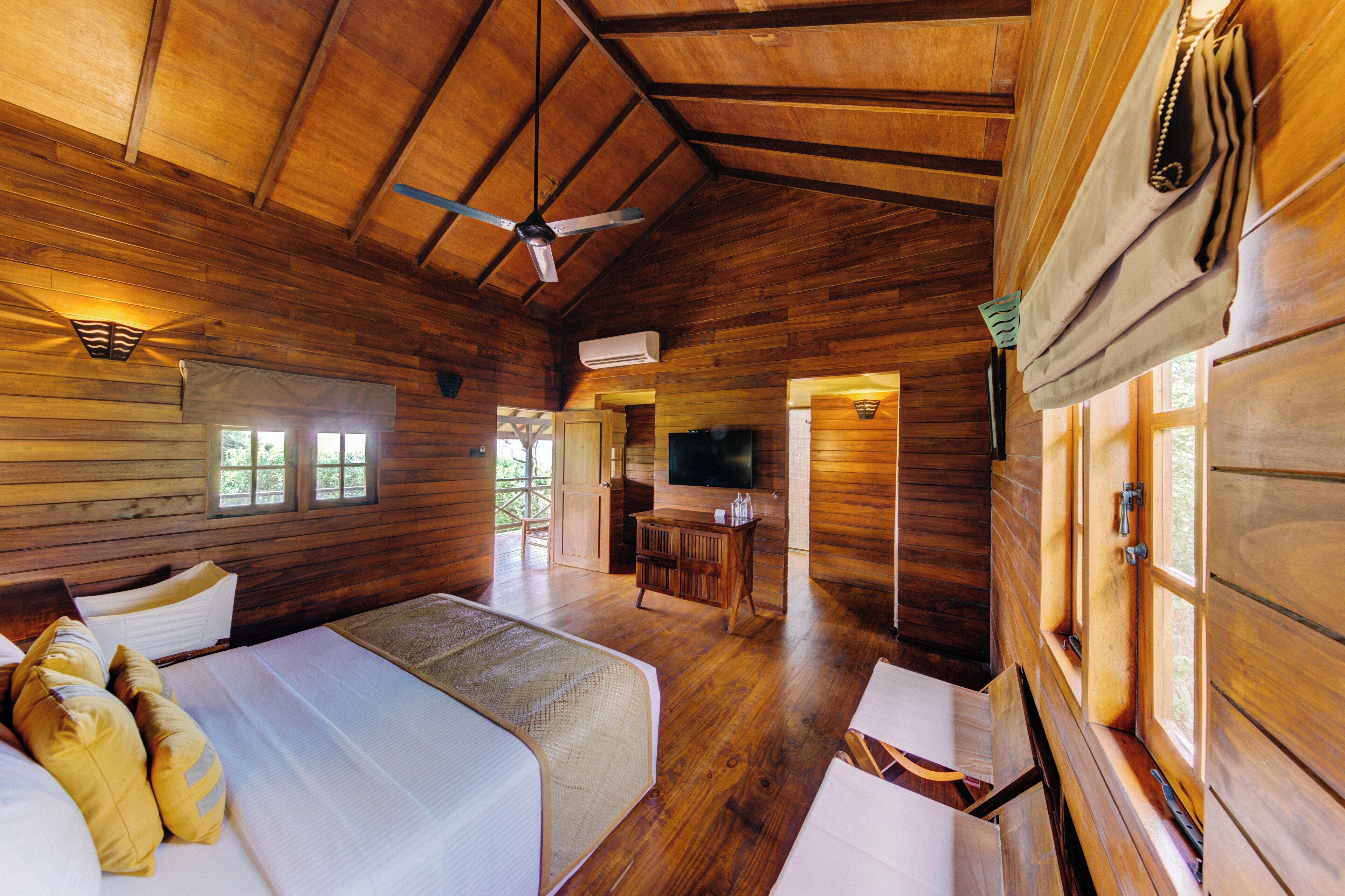 A wooden cabin room with two beds, a ceiling fan, a television, and a small kitchenette.