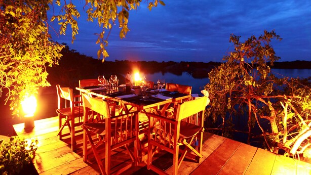 An outdoor dining area with a table set for two, surrounded by chairs, overlooking a body of water. - Cinnamon Wild Yala (Kirinda)