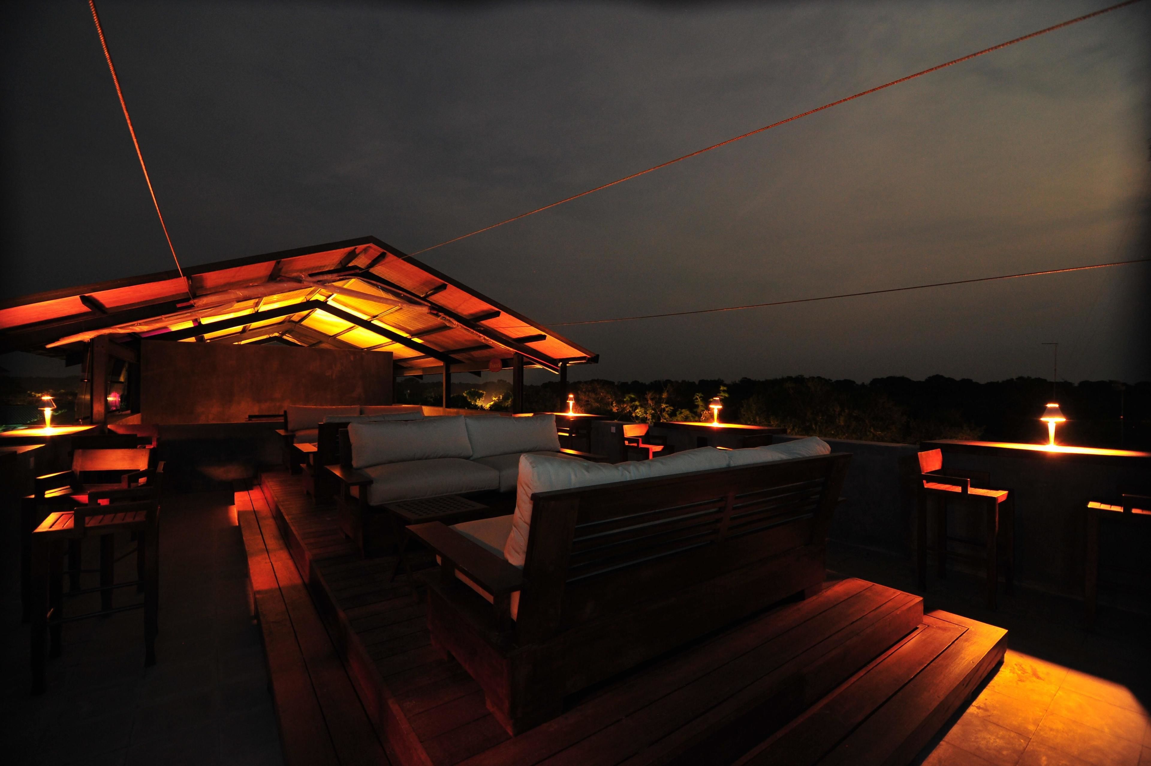 Terraza al aire libre con mobiliario de madera y una zona de estar techada, iluminada con luces cálidas.