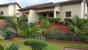 A residential building with a tiled roof, a stone retaining wall, and a well-maintained garden with various plants and a paved area. - Coconut Grove Miners Lodge (Obuasi)