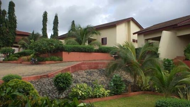 A residential area with a house featuring a sloped roof, a stone pathway, and a well-maintained garden with various plants and a palm tree. - Coconut Grove Miners Lodge (Obuasi)