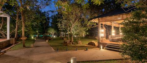 A pathway leading to a building with a covered outdoor area, surrounded by trees and illuminated by ambient lighting.