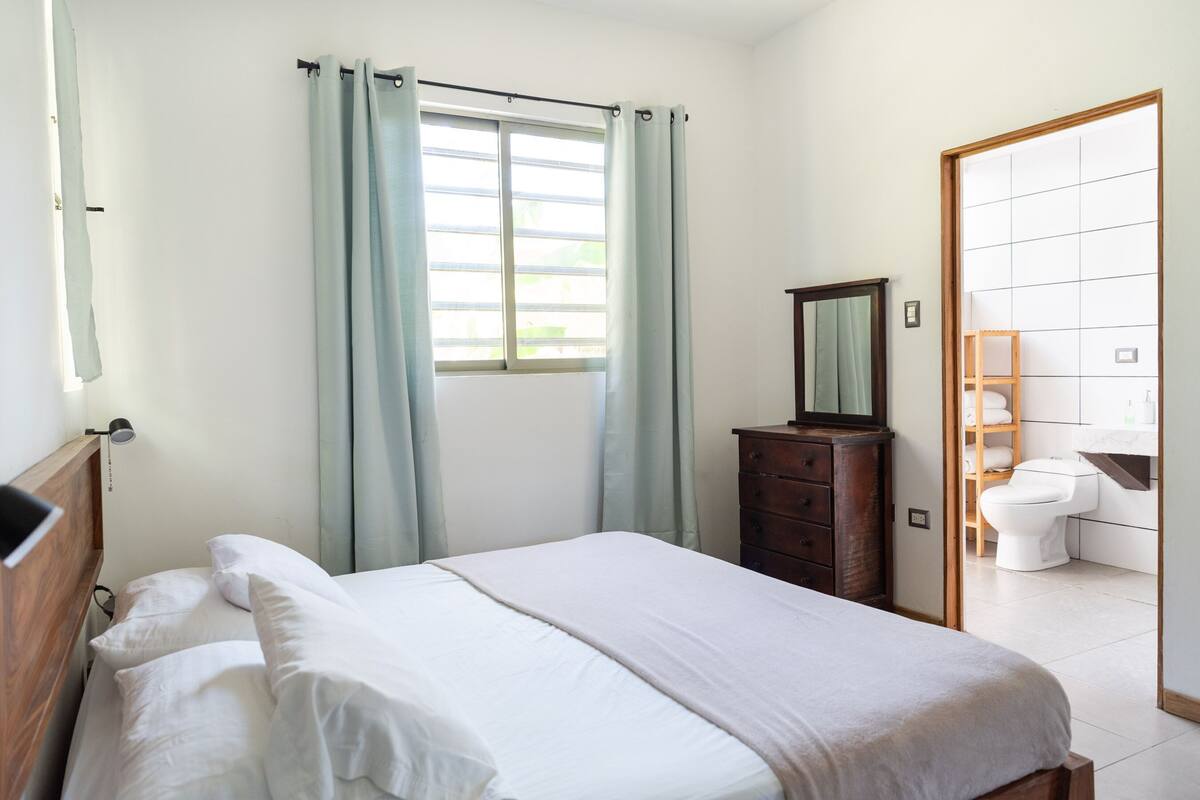 A bedroom with a bed, a wooden dresser, a mirror, and a view of the bathroom.