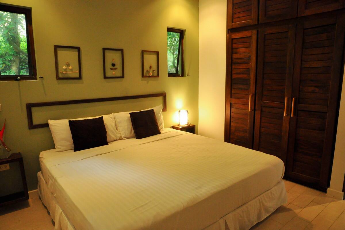 A neatly made bed with white and dark pillows, a bedside table with a lamp, framed pictures on the wall, and a wooden wardrobe.