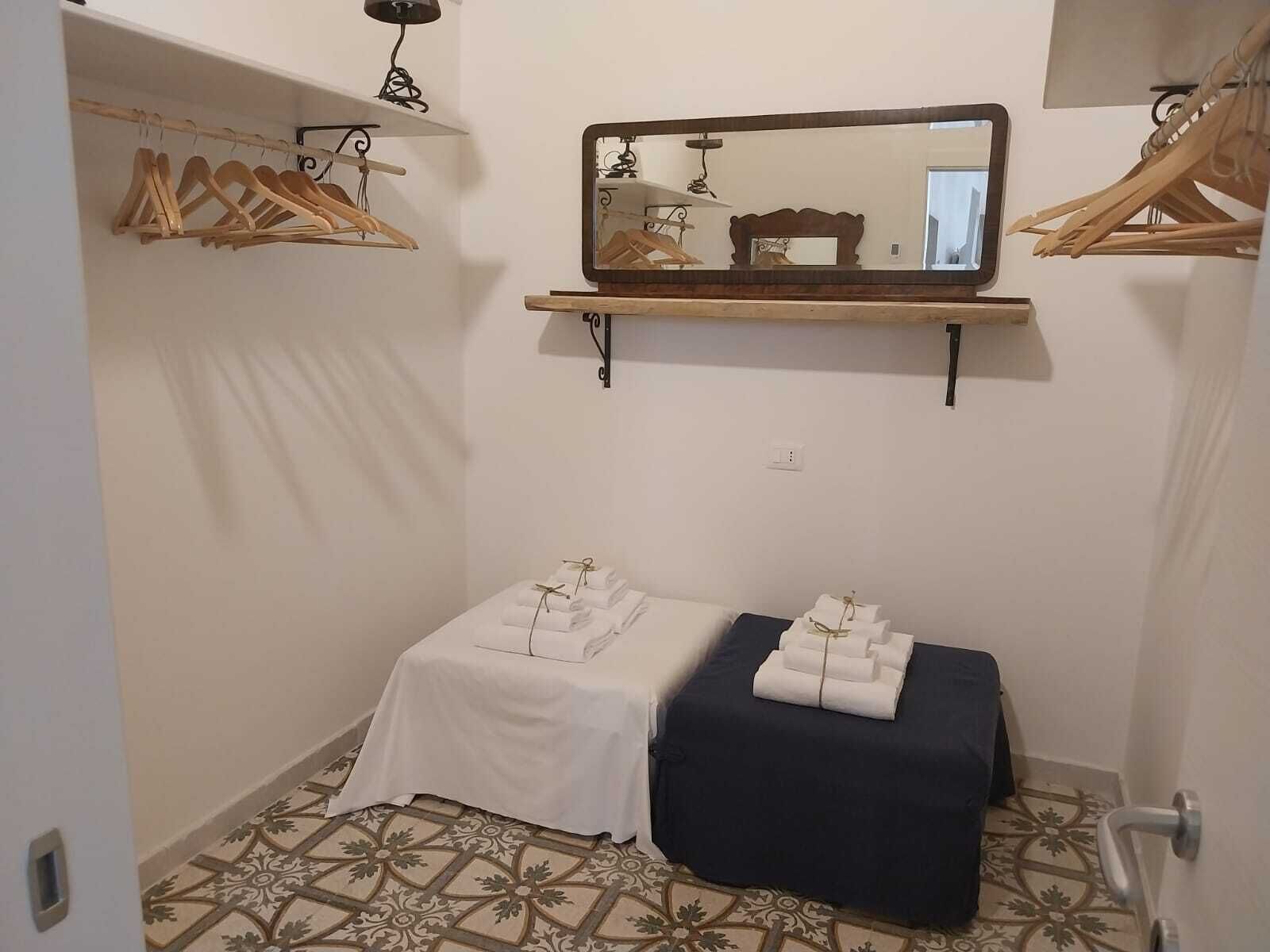 A small, clean room with a bed, a bench, a mirror, and wooden hangers on a shelf.