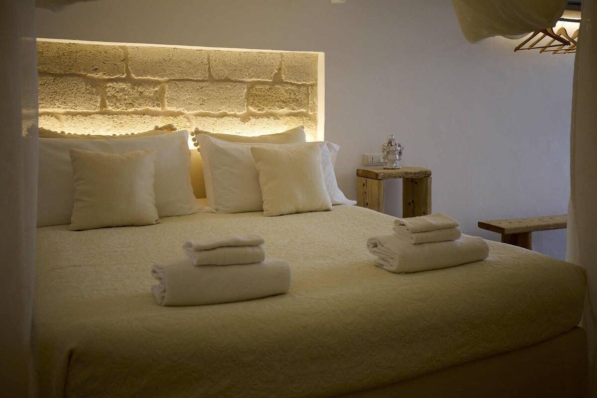 Un lit bien fait, avec du linge de lit blanc et des oreillers, une table de chevet agrémentée d’un objet décoratif et un luminaire fixé au mur.