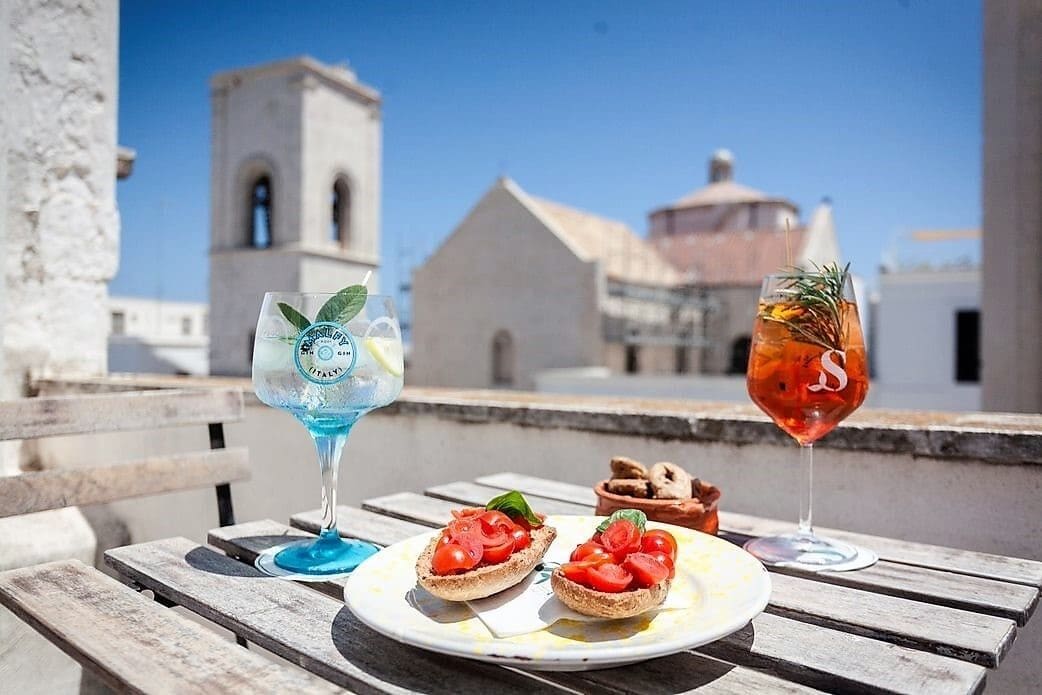 Una mesa con dos vasos de bebidas y un plato de bruschetta, frente a un edificio con una torre de campanas.