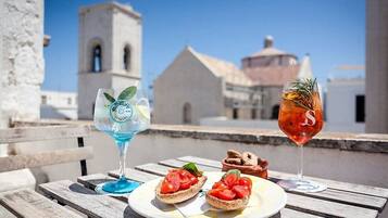 A table with two glasses of beverages and a plate with bruschetta, set against a backdrop of a building with a bell tower.