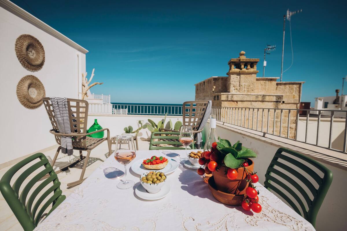 Une terrasse avec une table dressée pour un repas, des chaises, une vue sur la mer et un bâtiment en arrière-plan.