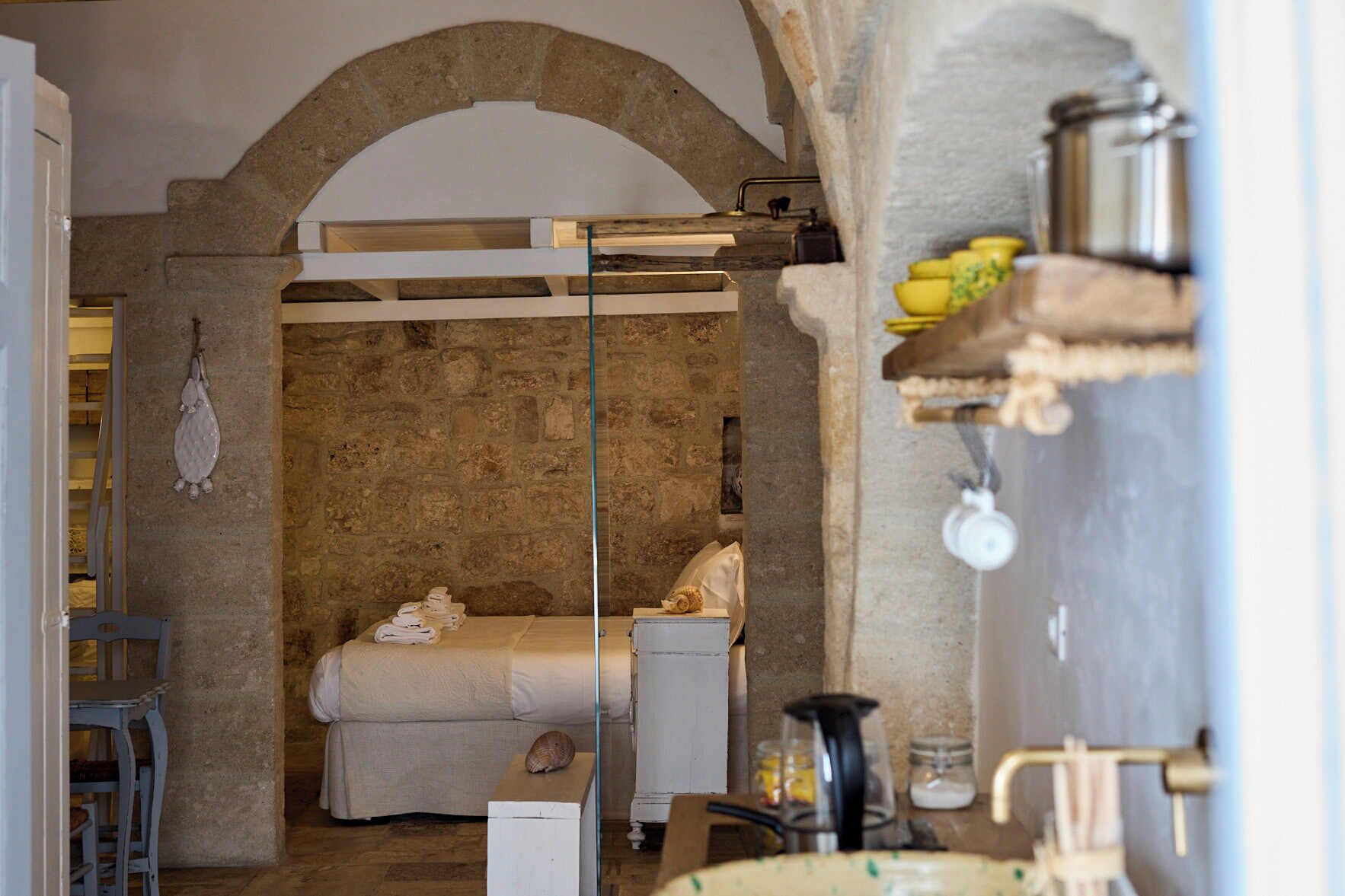 Une pièce rustique avec un mur en pierre, un lit avec un matelas blanc, une petite table ornée d’un coquillage et une étagère avec des bocaux et une bouilloire.