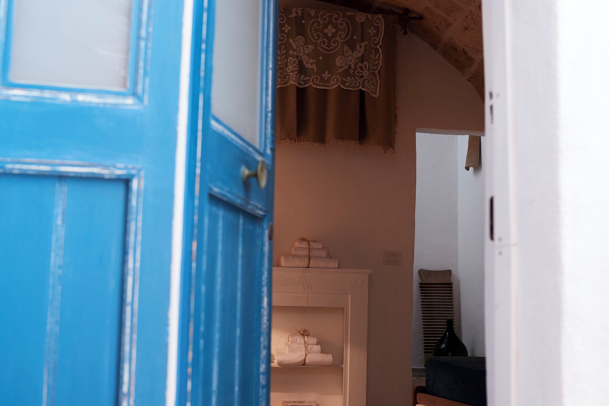 Une porte qui mène à une pièce où se trouve une étagère blanche, sur laquelle sont pliés des serviettes, et au-dessus de laquelle est accroché un rideau en dentelle.