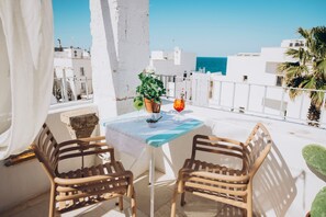 Een balkon met twee rieten stoelen, een kleine tafel met een bloempot, en uitzicht op de zee en witte gebouwen.