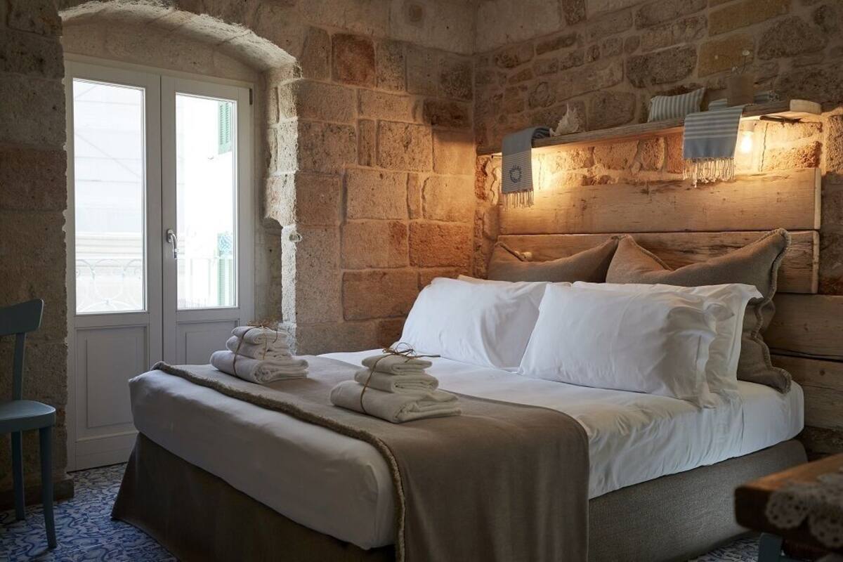 Une pièce aux murs de pierre, avec un lit, une chaise et une fenêtre.
