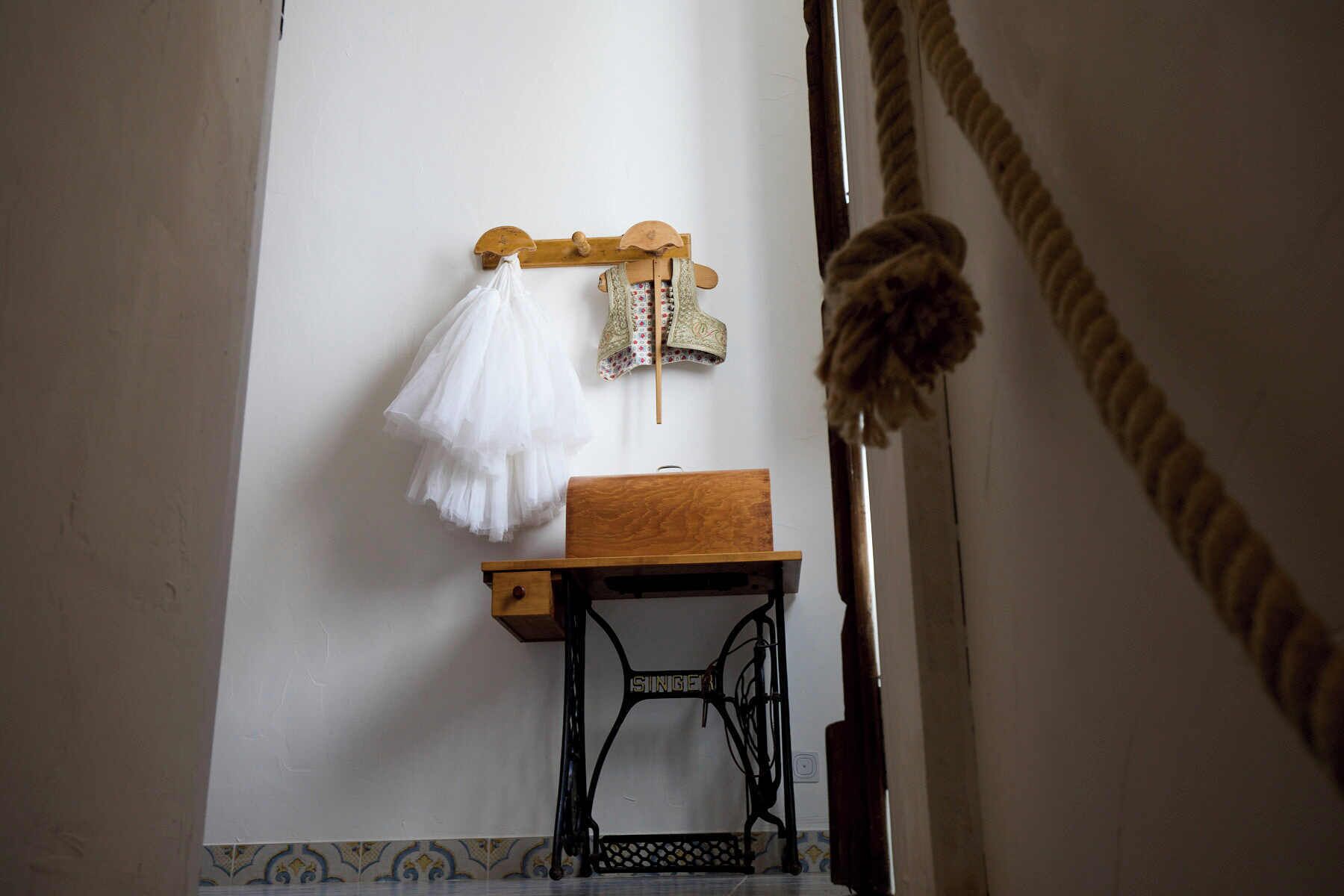 Une pièce avec une table en bois, une machine à coudre Singer et un porte-vêtements mural sur lequel sont suspendus un vêtement blanc et un gilet à motifs.