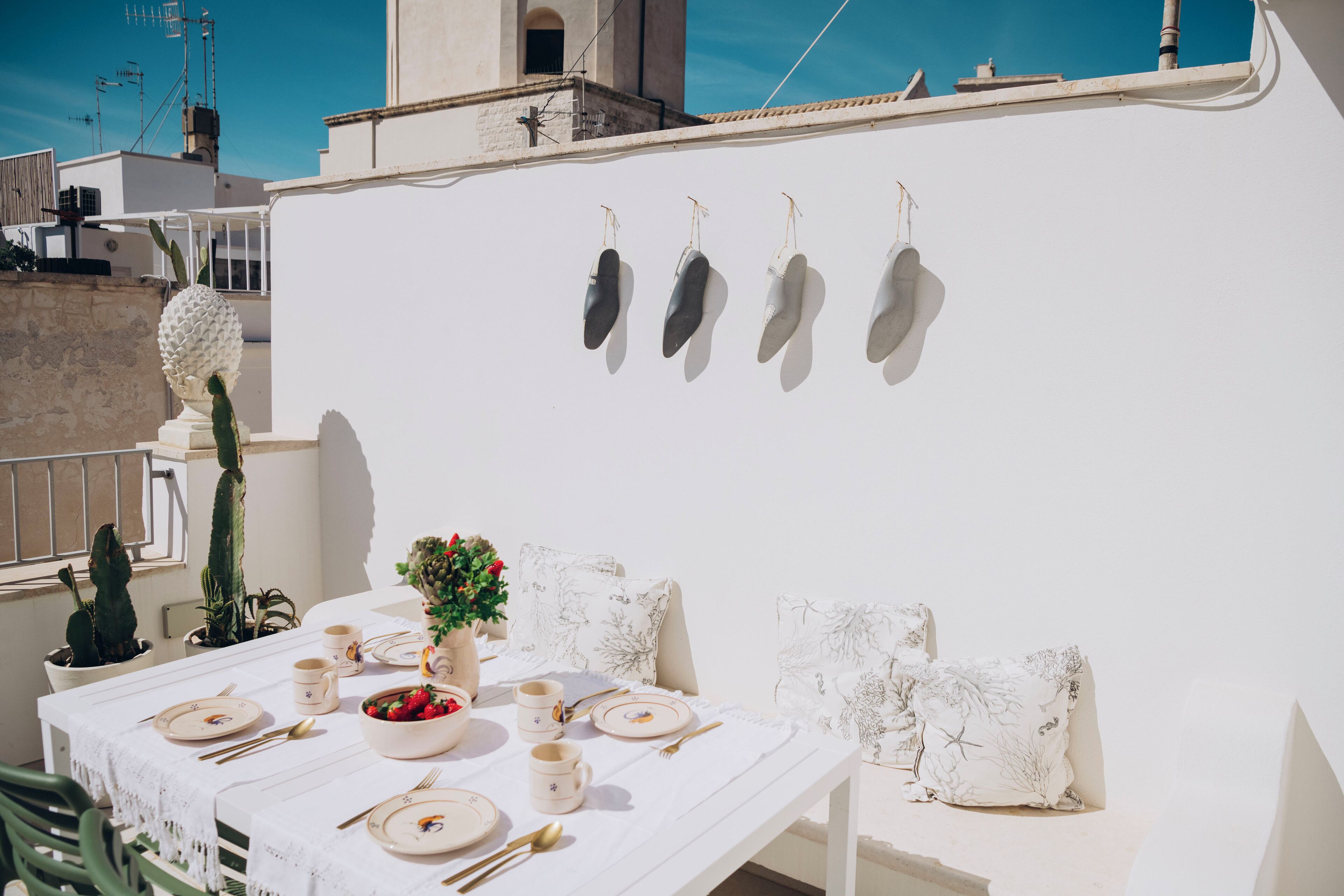 A white table set with plates, cutlery, and a bowl of fruit, against a white wall with a cactus and a hanging decoration.
