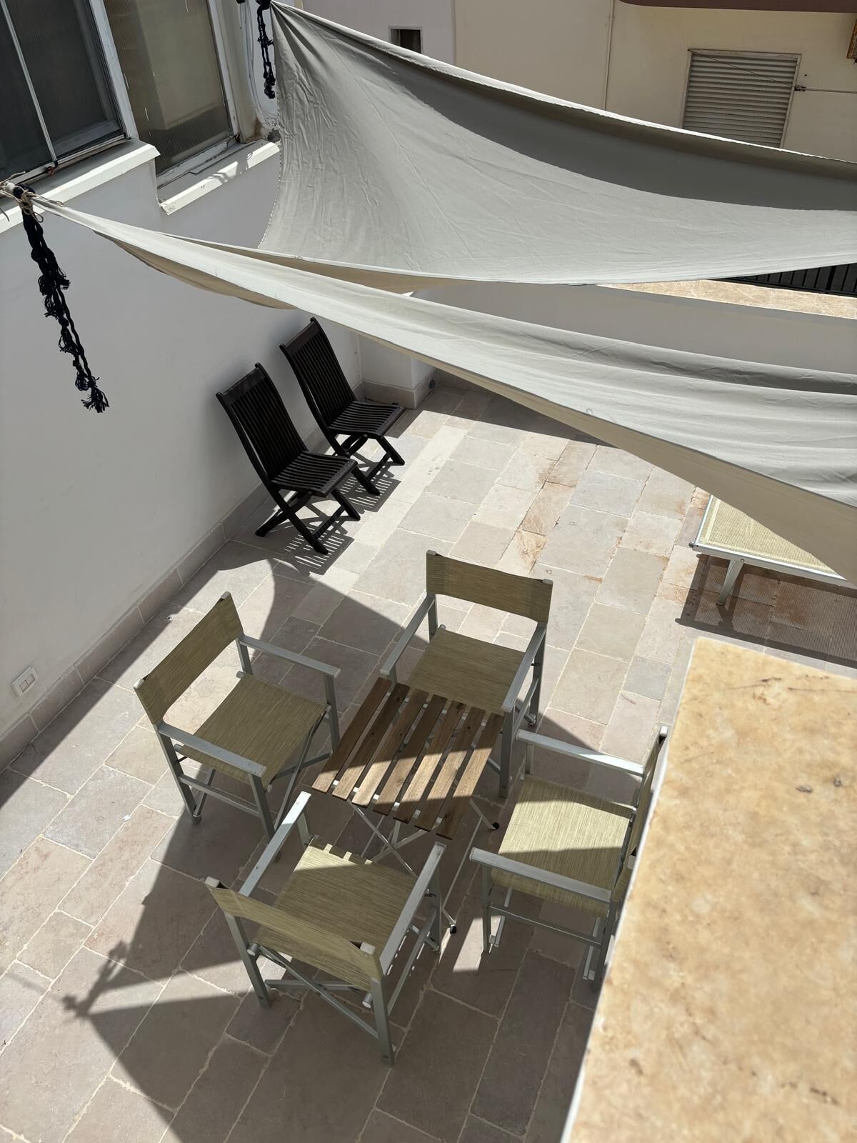 A patio with a table and chairs, a white shade sail, and a view of the building's interior.