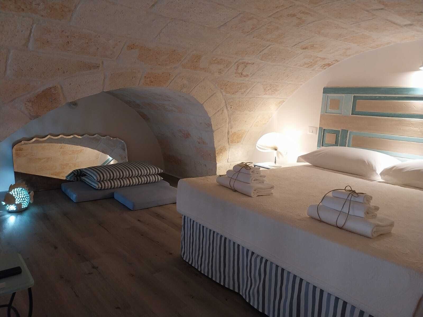 Um quarto aconchegante com parede de pedra, cama com lençóis brancos, abajur e uma pequena mesa com luminária.