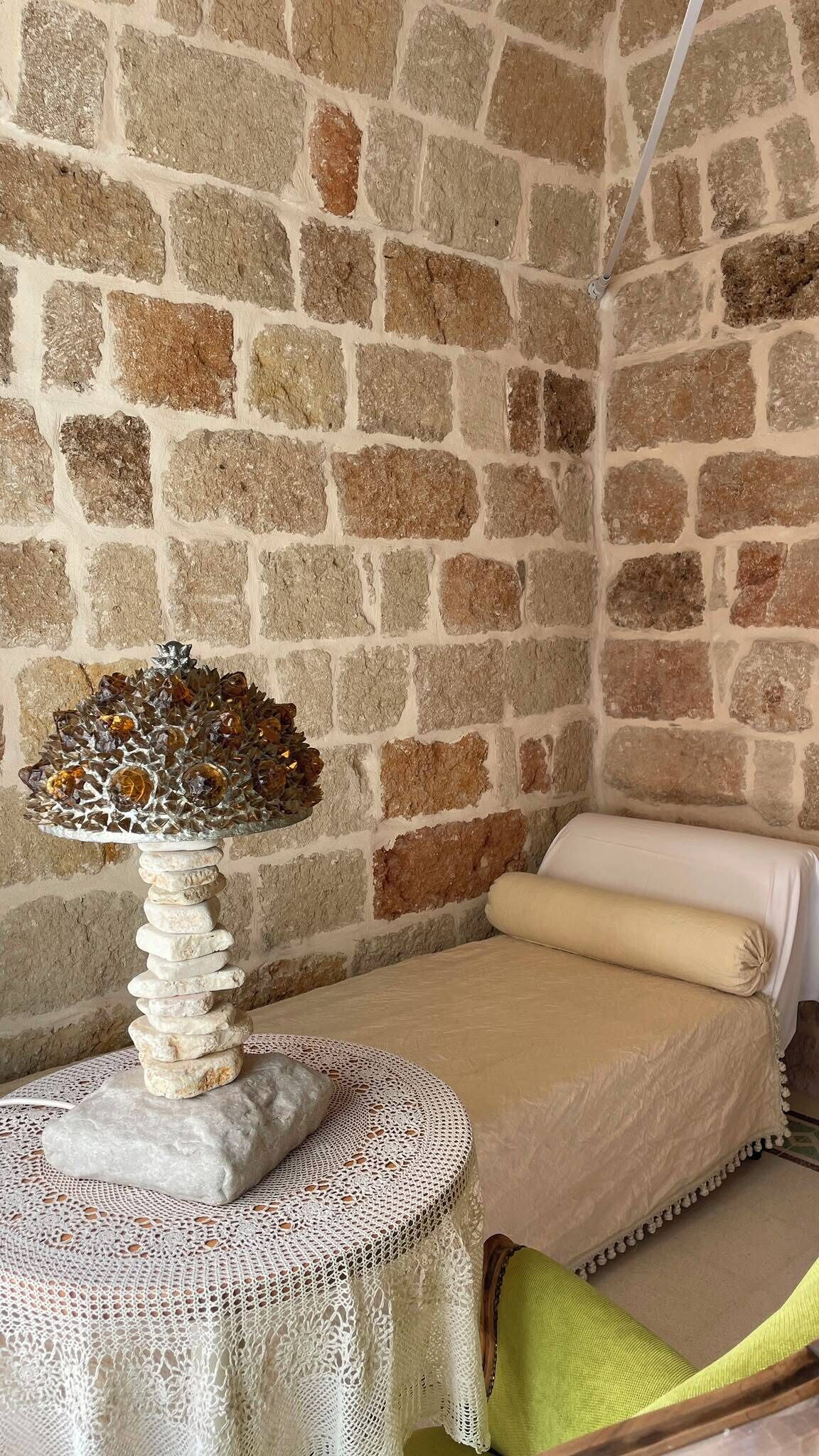 A room with a stone wall, a bed with a white cover, and a table with a decorative glass object and stones.