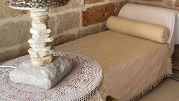 A room with a stone wall, a bed with a white cover, and a table with a decorative glass object and stones.
