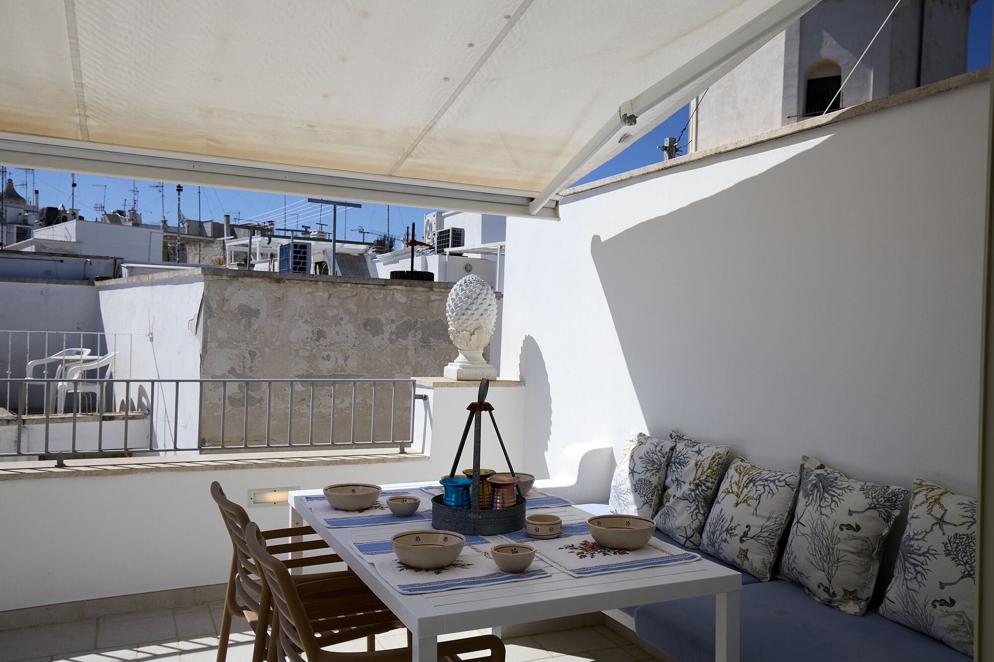 A balcony with a table set for dining, a white wall, and a view of buildings in the background.