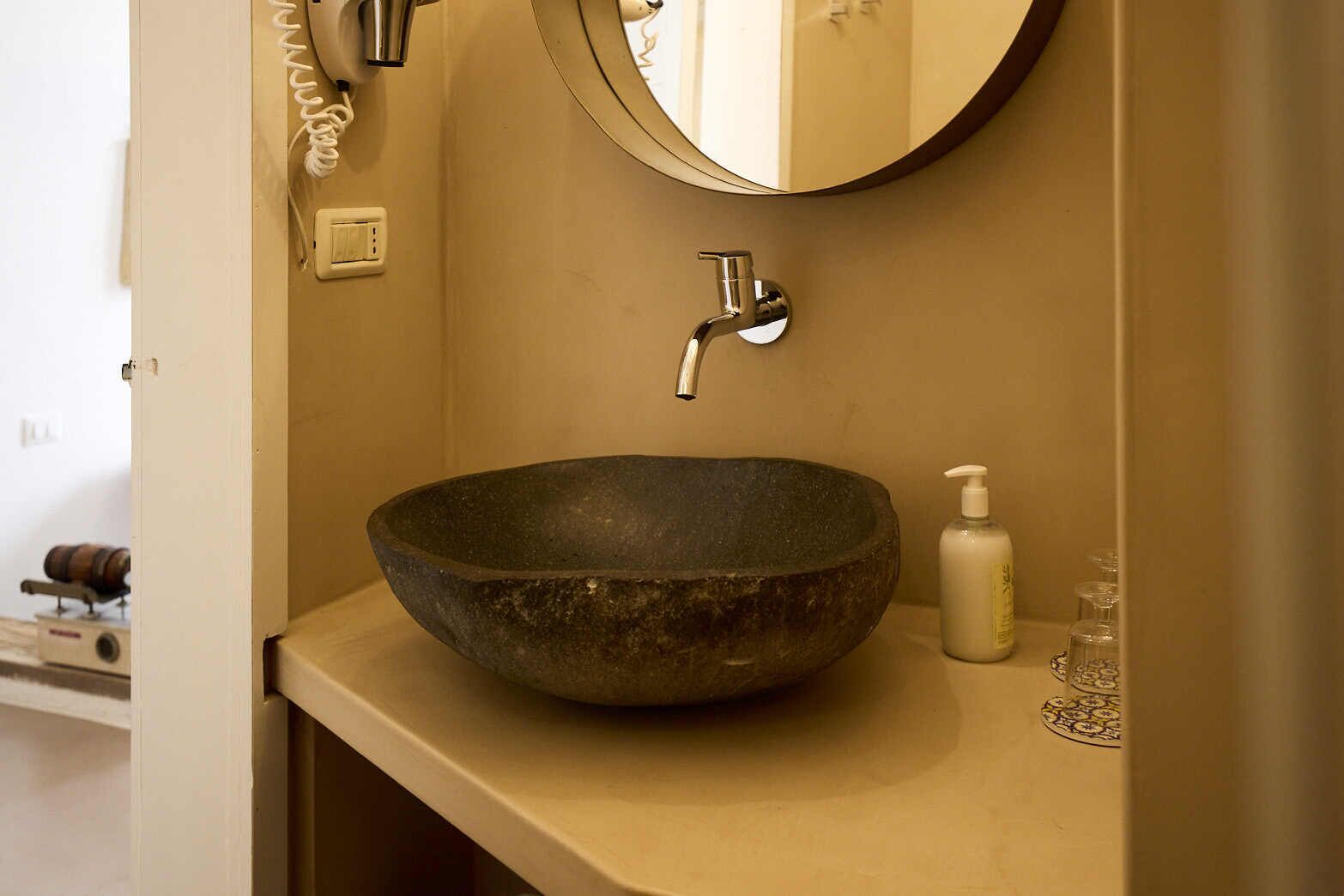 A bathroom with a large, dark stone sink, a modern faucet, a round mirror, and a small bottle of soap on the counter.