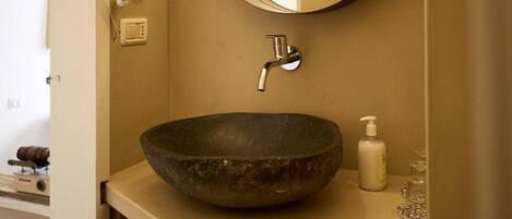 Un baño con un lavabo grande de piedra oscura, un grifo moderno, un espejo redondo y una pequeña botella de jabón en la encimera.