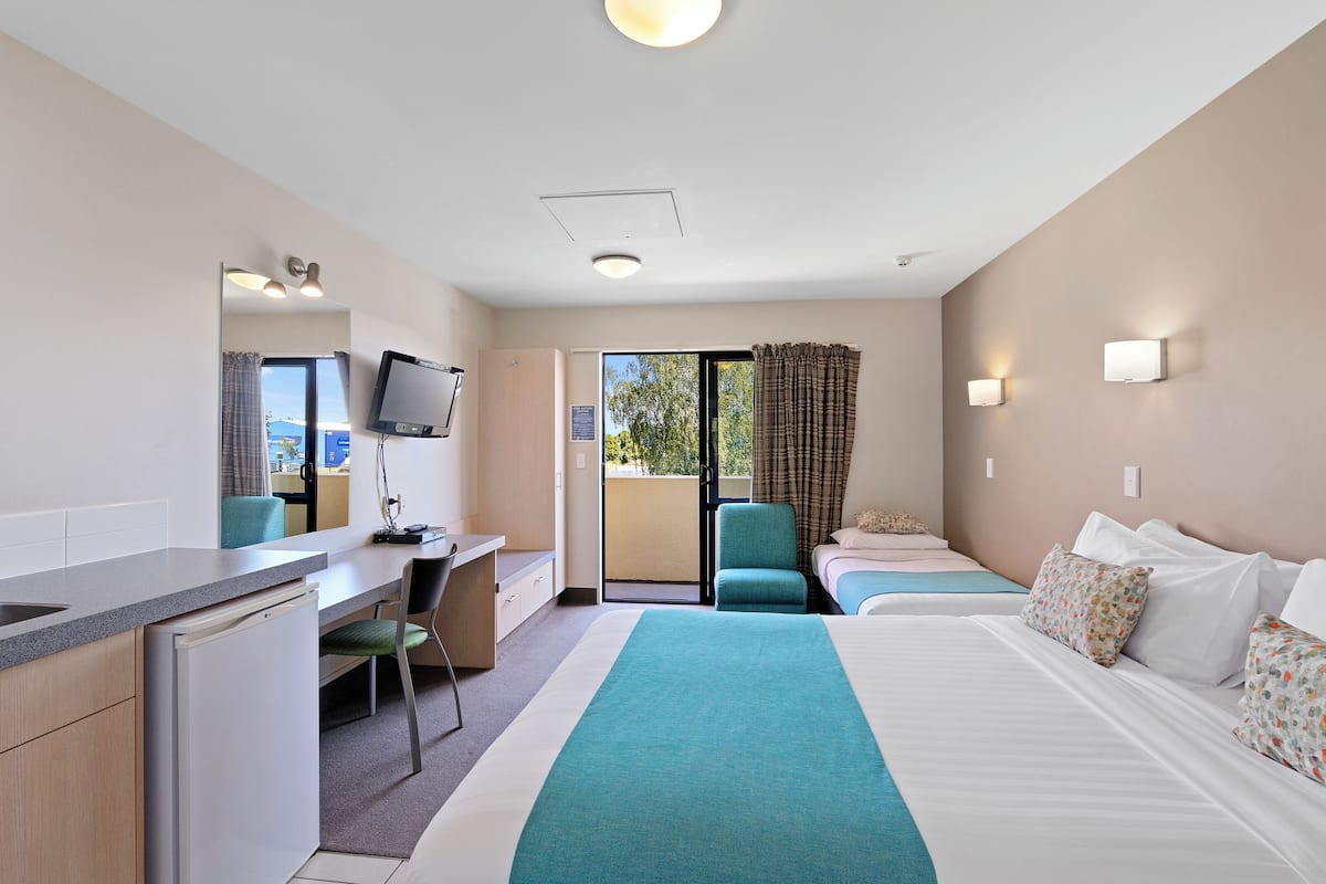 Habitación de hotel con cama, escritorio, silla, televisor y balcón con cortinas.